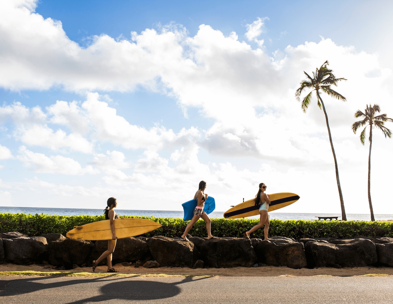 3 people with various surf boards walking along beachside